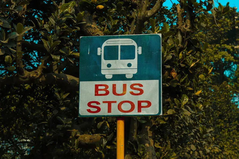 The stop sign carries only a pictogram and the words "BUS STOP" — no place name, no route number — so you identify your bus from the Bengali board on its front.