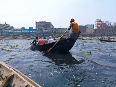 A hand-rowed ferry crossing the Buriganga — the boatman stands and rows at the stern while passengers sit low in the hull