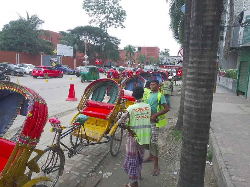 Rickshaws lined up along the kerb waiting for fares. The numbers on the vests are a local neighbourhood association's registration, not a city-wide scheme.