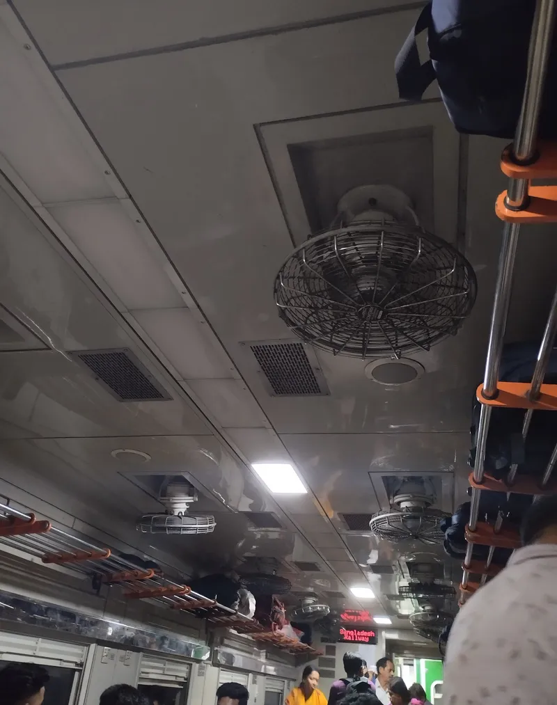 The ceiling of a non-air-conditioned coach — rotating fans, steel luggage racks, and standing passengers in the aisle