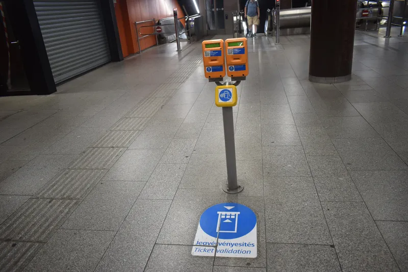 Validators at Corvin-negyed station. There are no fare gates: a paper ticket goes into the slot on the orange unit, the yellow pad below is an NFC point for a phone, and the blue disc on the floor reads "Jegyérvényesítés / Ticket validation".