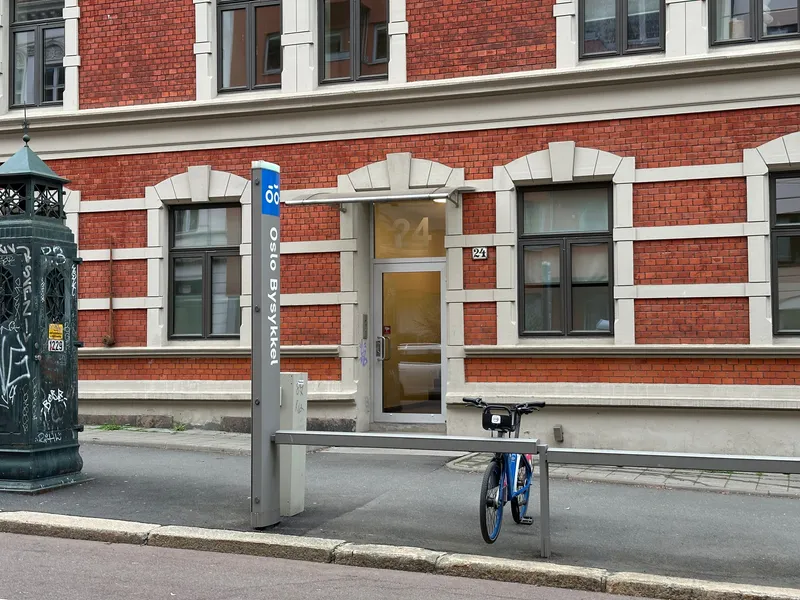A station is marked by a post reading "Oslo Bysykkel" - grey, with a blue panel at the top. Bikes lock into the rack beside it by the front wheel.