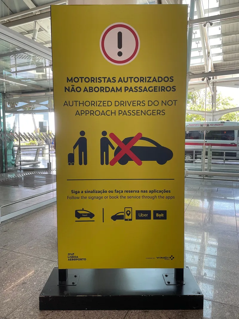 Notice in Terminal 1 at Lisbon Airport: "Authorized drivers do not approach passengers - follow the signage or book the service through the apps", with taxi, Uber and Bolt logos.